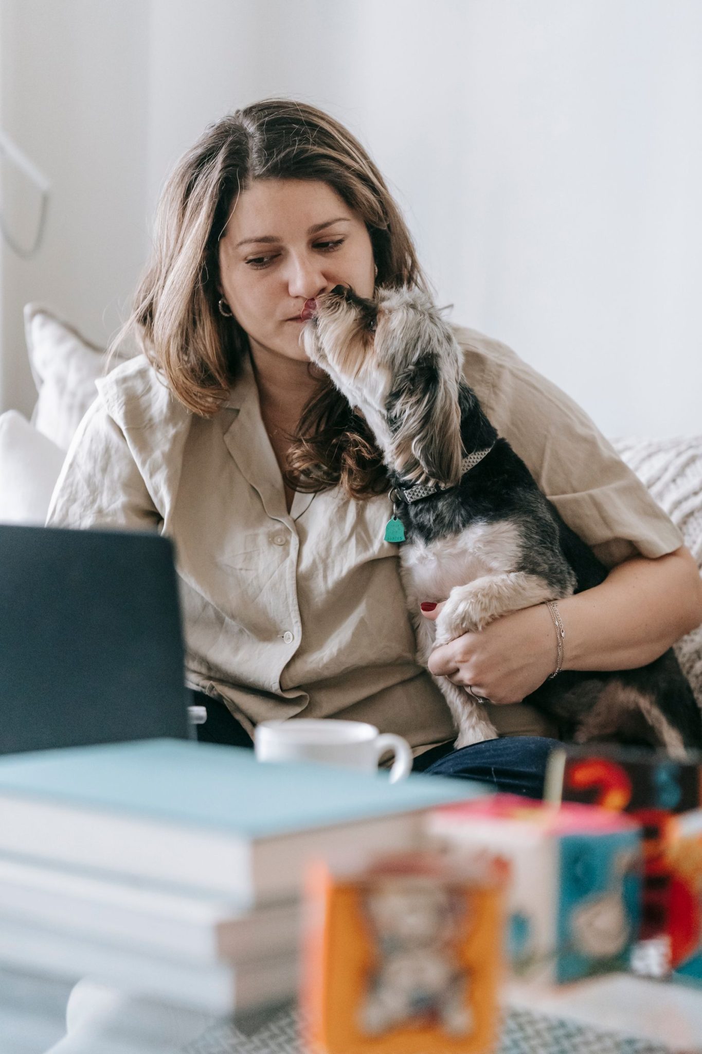 Kleine hond die het gezicht likt van zijn eigenaar, die achter een laptop aan tafel zit met papieren.